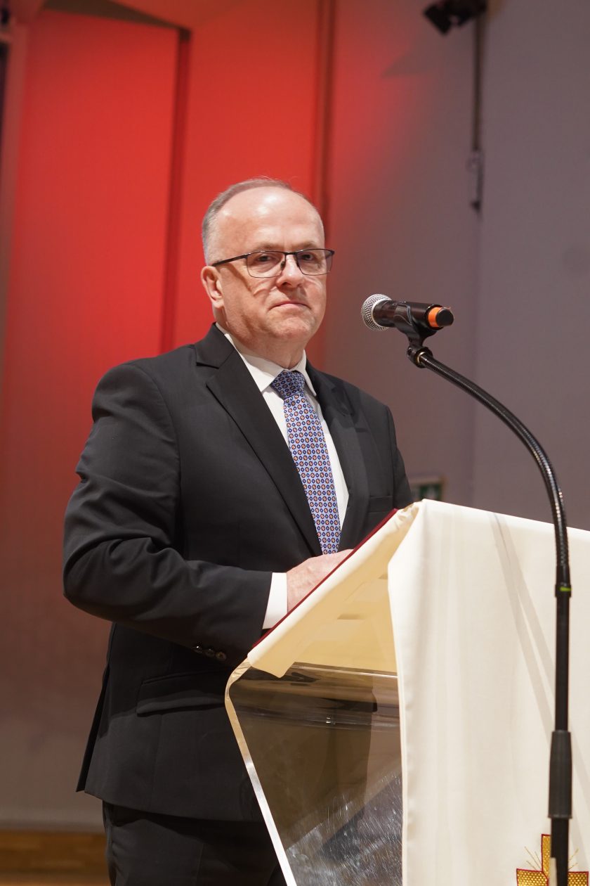 Zdjęcie przedstawia prezesa Bonifraterskiego Centrum Medycznego stojącego przy mównicy podczas uroczystej gali inauguracji nowej odsłony Szpitala Zakonu Bonifratrów pw. Aniołów Stróżów w Katowicach. Mężczyzna ubrany jest w ciemny garnitur, białą koszulę i wzorzysty krawat. Przemawia do zgromadzonych gości przy mikrofonie ustawionym na mównicy przykrytej białym obrusem z haftowanym krzyżem. W tle widoczne są czerwone elementy oświetlenia scenicznego