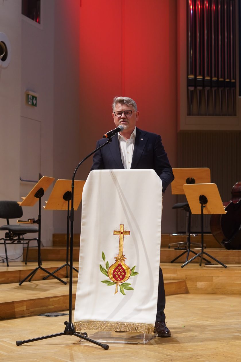 Zdjęcie przedstawia przedstawiciela Bonifraterskiego Centrum Medycznego przemawiającego z mównicy podczas uroczystej gali inaugurującej nową odsłonę Szpitala Zakonu Bonifratrów pw. Aniołów Stróżów w Katowicach. Mężczyzna ubrany jest w granatowy garnitur i białą koszulę. Stoi za przezroczystą mównicą przykrytą białym obrusem z haftowanym symbolem krzyża i owocu granatu. W tle widoczne są pulpity dla muzyków oraz instrumenty przygotowane do koncertu, a ciepłe oświetlenie sceniczne nadaje wnętrzu elegancki i uroczysty charakter