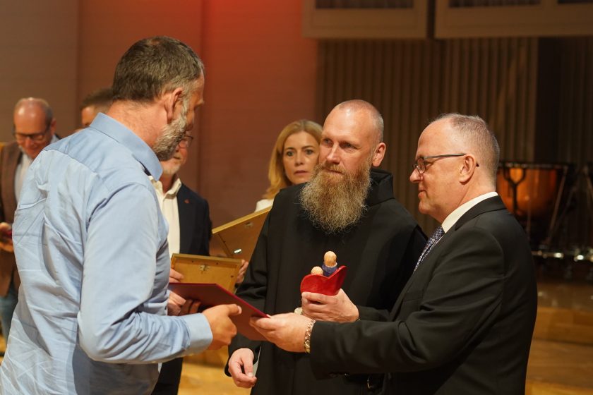 Zdjęcie przedstawia moment wręczenia pamiątkowej statuetki podczas uroczystej gali inaugurującej nową odsłonę Szpitala Zakonu Bonifratrów pw. Aniołów Stróżów w Katowicach. W centrum kadru stoi zakonnik Zakonu Bonifratrów w czarnym habicie z charakterystyczną długą brodą, który trzyma ceramiczną statuetkę przedstawiającą dwie stylizowane postacie z czerwonym sercem. Obok niego znajduje się mężczyzna w ciemnym garniturze i okularach wręczający laureatowi dyplom, natomiast po lewej stronie odbiorca nagrody odbiera dokument. W tle widoczni są pozostali uczestnicy uroczystości, trzymający dyplomy i statuetki. Całość odbywa się na scenie sali koncertowej oświetlonej ciepłym światłem
