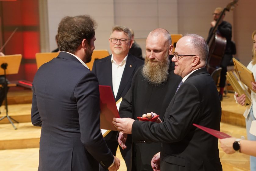 Zdjęcie przedstawia moment wręczenia pamiątkowego dyplomu podczas gali inaugurującej nową odsłonę Szpitala Zakonu Bonifratrów pw. Aniołów Stróżów w Katowicach. W centrum kadru przedstawiciel Bonifraterskiego Centrum Medycznego w ciemnym garniturze przekazuje czerwony dyplom mężczyźnie stojącemu tyłem do obiektywu. Obok znajduje się zakonnik Zakonu Bonifratrów z długą brodą, trzymający ceramiczną statuetkę z motywem czerwonego serca. W tle widoczni są pozostali uczestnicy uroczystości oraz muzycy przygotowani do koncertu