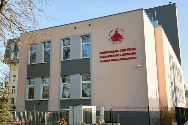 Widok na budynek Regionalnego Centrum Diagnostyki i Leczenia Chorób Onkologicznych im. Hanny Olszewskiej. Na jasnej elewacji umieszczono czerwone logo oraz nazwę placówki. Obiekt ma nowoczesną architekturę z dużymi prostokątnymi oknami i szaro-beżową kolorystyką. W kadrze widoczne są także ogrodzenie, fragmenty roślinności oraz drzewa otaczające budynek