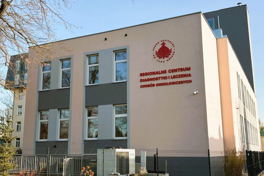 Widok na budynek Regionalnego Centrum Diagnostyki i Leczenia Chorób Onkologicznych im. Hanny Olszewskiej. Na jasnej elewacji umieszczono czerwone logo oraz nazwę placówki. Obiekt ma nowoczesną architekturę z dużymi prostokątnymi oknami i szaro-beżową kolorystyką. W kadrze widoczne są także ogrodzenie, fragmenty roślinności oraz drzewa otaczające budynek