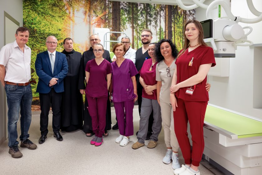 Zdjęcie przedstawia wspólne pamiątkowe zdjęcie pracowników Pracowni Diagnostyki Obrazowej, przedstawicieli Zakonu Bonifratrów oraz władz szpitala. Grupa stoi obok nowoczesnego aparatu RTG. Pracownicy medyczni ubrani są w kolorowe uniformy, a w tle znajduje się fototapeta przedstawiająca słoneczny las. Po prawej stronie widoczny jest stół diagnostyczny oraz elementy aparatu RTG. Wszyscy uczestnicy patrzą w stronę obiektywu i uśmiechają się.