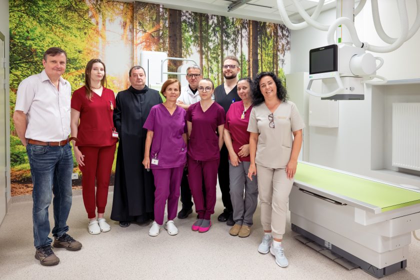 Zdjęcie przedstawia drugie pamiątkowe zdjęcie zespołu Pracowni Diagnostyki Obrazowej wykonane po zakończeniu uroczystości. W centrum stoją pracownicy medyczni w fioletowych, bordowych, szarych i białych uniformach oraz bonifrater w czarnym habicie. Obok nich znajdują się przedstawiciele szpitala. Po prawej stronie widoczny jest nowoczesny aparat RTG wraz ze stołem diagnostycznym, a tło stanowi dekoracyjna fototapeta przedstawiająca las.