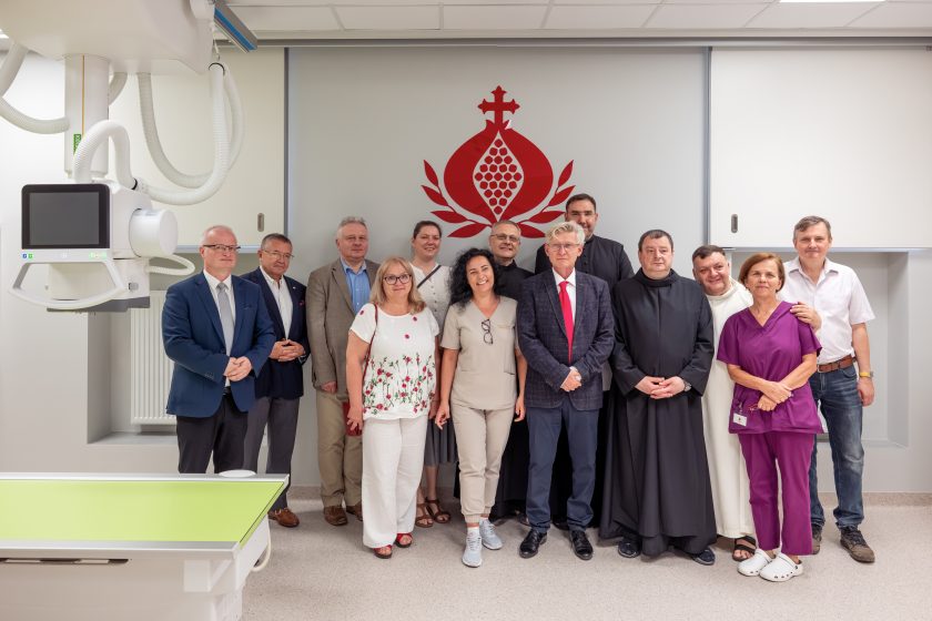 Zdjęcie przedstawia wspólne zdjęcie uczestników uroczystości poświęcenia zmodernizowanej Pracowni Diagnostyki Obrazowej. W pierwszym rzędzie stoją przedstawiciele władz szpitala, pracownicy medyczni oraz bonifratrzy, a za nimi pozostali uczestnicy wydarzenia. Po lewej stronie widoczny jest fragment nowoczesnego aparatu RTG, natomiast na ścianie za grupą znajduje się duże czerwone logo Zakonu Bonifratrów. Zdjęcie wykonano we wnętrzu nowoczesnej pracowni diagnostycznej.