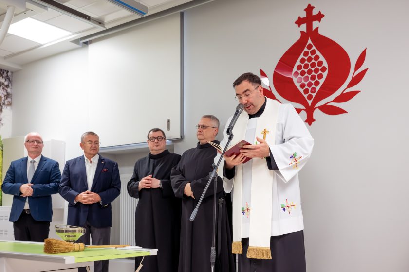 Zdjęcie przedstawia uroczystość poświęcenia nowo wyremontowanej Pracowni Diagnostyki Obrazowej w Szpitalu Zakonu Bonifratrów św. Jana Grandego w Krakowie. Na pierwszym planie kapłan ubrany w białą stułę stoi przy mikrofonie i odczytuje modlitwy z czerwonej księgi. Obok niego stoją dwaj bonifratrzy w czarnych habitach oraz dwóch przedstawicieli władz szpitala ubranych w garnitury. Na zielonym blacie stołu znajdują się kropidło oraz misa z wodą święconą przygotowane do obrzędu poświęcenia. W tle widoczne jest duże czerwone logo Zakonu Bonifratrów umieszczone na jasnoszarej ścianie. Wnętrze pracowni jest nowoczesne, jasne i przestronne.