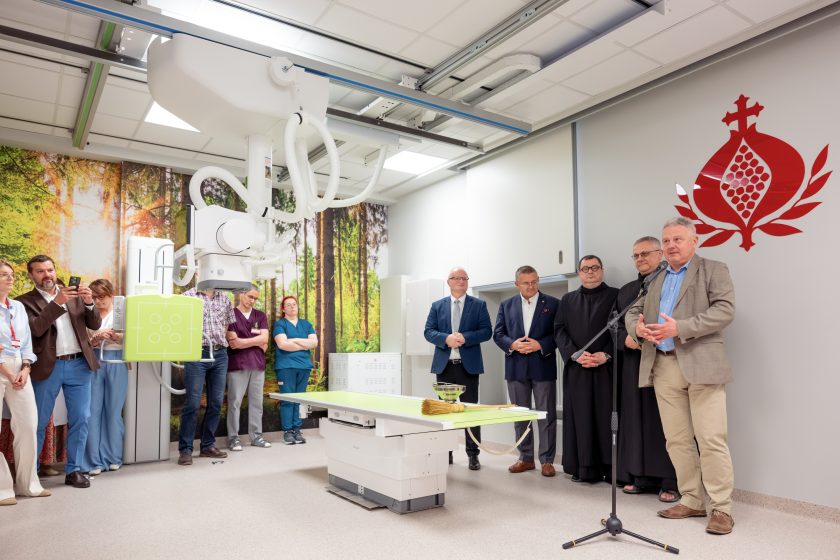 Zdjęcie przedstawia przemówienie podczas uroczystego otwarcia zmodernizowanej Pracowni Diagnostyki Obrazowej. Na pierwszym planie znajduje się nowoczesny aparat RTG z zawieszeniem sufitowym oraz stół diagnostyczny z zielonym blatem. Przy mikrofonie przemawia mężczyzna w jasnej marynarce, obok stoją przedstawiciele szpitala i Zakonu Bonifratrów. Po lewej stronie zgromadzili się pracownicy medyczni oraz zaproszeni goście obserwujący wydarzenie. Ścianę pracowni zdobi fototapeta przedstawiająca leśny krajobraz, a po prawej stronie znajduje się czerwone logo Zakonu Bonifratrów. Na stole leżą misa z wodą święconą i kropidło przygotowane do poświęcenia pracowni.