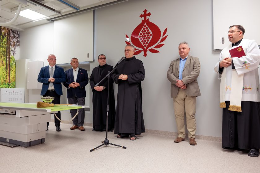 Zdjęcie przedstawia przemówienie jednego z bonifratrów podczas uroczystości otwarcia nowej pracowni RTG. Duchowny w czarnym habicie stoi przy mikrofonie i zwraca się do zgromadzonych gości. Za nim stoją przedstawiciele władz szpitala, drugi bonifrater oraz kapłan ubrany w białą stułę trzymający księgę liturgiczną. Po lewej stronie widoczny jest fragment nowoczesnego stołu diagnostycznego oraz misa z wodą święconą i kropidło. W tle znajduje się charakterystyczne czerwone logo Zakonu Bonifratrów na jasnej ścianie.