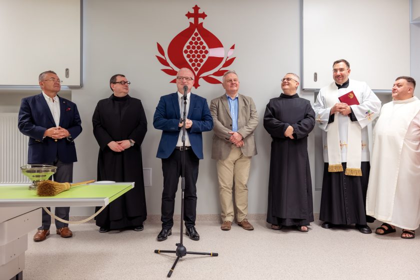 Zdjęcie przedstawia przemówienie dyrektora szpitala podczas uroczystości otwarcia Pracowni Diagnostyki Obrazowej. Dyrektor stoi przy mikrofonie i przemawia do zgromadzonych gości. Obok niego stoją przedstawiciele Zakonu Bonifratrów, duchowny prowadzący uroczystość oraz zaproszeni goście. Na pierwszym planie widoczne są misa z wodą święconą i kropidło. Nad uczestnikami znajduje się czerwone logo Zakonu Bonifratrów umieszczone na ścianie. Na twarzach zgromadzonych widoczne są uśmiechy i życzliwa atmosfera.