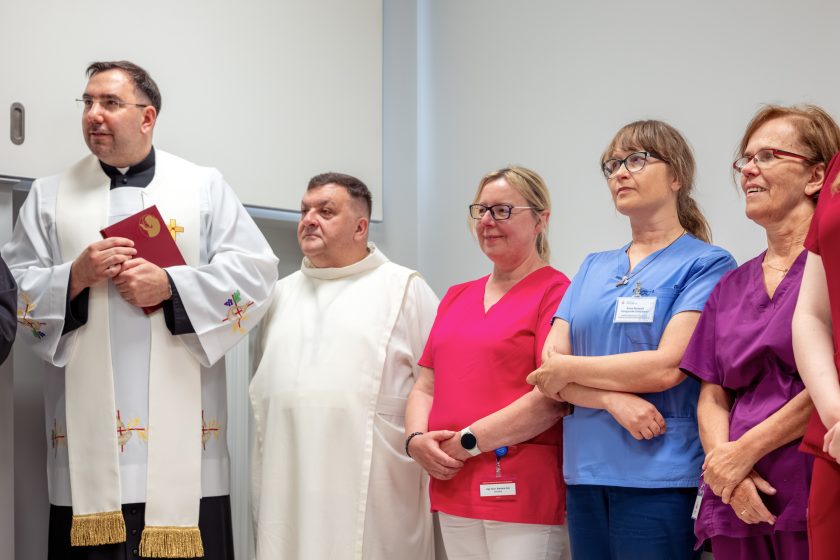 Zdjęcie przedstawia kapłana prowadzącego uroczystość oraz pracowników medycznych zgromadzonych podczas poświęcenia nowej pracowni RTG. Po lewej stronie stoi duchowny ubrany w białą stułę, obok niego bonifrater w białym habicie. Dalej ustawione są pracownice personelu medycznego w różowych, niebieskich i fioletowych uniformach, uważnie słuchające przebiegu uroczystości. Wszyscy stoją na tle jasnej ściany w nowoczesnym wnętrzu pracowni.