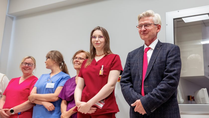 Zdjęcie przedstawia grupę pracowników medycznych oraz jednego z gości honorowych uczestniczących w uroczystości otwarcia pracowni. Po prawej stronie stoi mężczyzna w eleganckim garniturze z czerwonym krawatem, obok niego pracownice szpitala ubrane w kolorowe uniformy medyczne. W centrum znajduje się młoda pracownica w bordowym stroju medycznym z identyfikatorem. Wszyscy stoją w jasnym pomieszczeniu diagnostycznym i z uwagą obserwują przebieg wydarzenia.