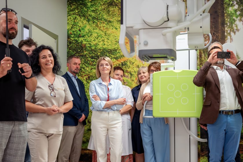 Zdjęcie przedstawia uczestników uroczystości zgromadzonych wokół nowoczesnego aparatu RTG podczas oficjalnego otwarcia pracowni. Na pierwszym planie widoczna jest część urządzenia wraz z zielonym panelem do wykonywania badań. W tle stoją pracownicy szpitala oraz zaproszeni goście obserwujący przebieg wydarzenia. Po prawej stronie mężczyzna w brązowej marynarce wykonuje zdjęcie telefonem komórkowym. Ścianę za uczestnikami zdobi fototapeta przedstawiająca leśny krajobraz, która tworzy przyjazne tło dla nowoczesnej aparatury diagnostycznej.