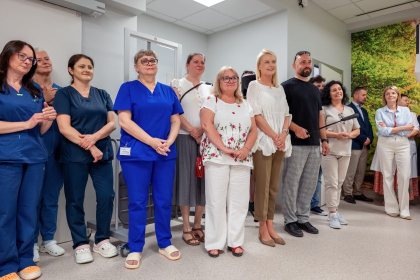 Zdjęcie przedstawia grupę pracowników medycznych oraz zaproszonych gości uczestniczących w uroczystości otwarcia nowej Pracowni Diagnostyki Obrazowej. W pierwszym rzędzie stoją osoby ubrane zarówno w stroje medyczne w odcieniach granatu i niebieskiego, jak i eleganckie ubrania. Wśród uczestników znajduje się także mężczyzna trzymający wysięgnik z mikrofonem. Wszyscy z uwagą obserwują przebieg wydarzenia. W tle widoczna jest część pracowni oraz fragment fototapety przedstawiającej las.
