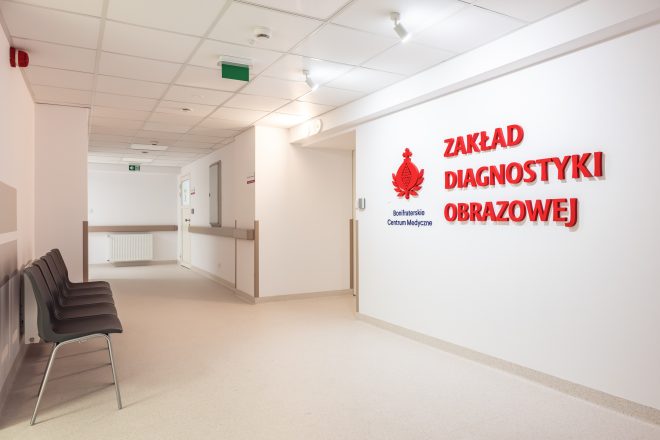 Zdjęcie przedstawia korytarz prowadzący do Zakładu Diagnostyki Obrazowej po zakończonej modernizacji. Po prawej stronie na białej ścianie umieszczono przestrzenny czerwony napis „Zakład Diagnostyki Obrazowej” wraz z logo Bonifraterskiego Centrum Medycznego. Po lewej stronie znajduje się rząd krzeseł przeznaczonych dla oczekujących pacjentów. Jasne ściany, nowoczesne oświetlenie oraz przestronny układ korytarza podkreślają estetyczny i funkcjonalny charakter wyremontowanej części szpitala.