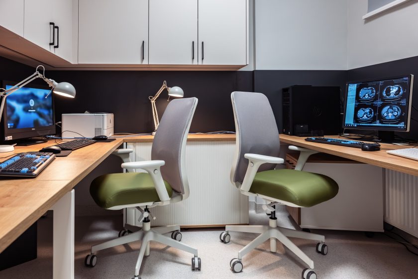 Zdjęcie przedstawia nowoczesne stanowiska opisowe w Zakładzie Diagnostyki Obrazowej. W pomieszczeniu znajdują się dwa ergonomiczne fotele z jasnozielonymi siedziskami ustawione przy drewnianych biurkach wyposażonych w komputery, monitory diagnostyczne oraz lampki biurkowe. Na jednym z monitorów wyświetlane są obrazy tomografii komputerowej. Nad stanowiskami zamontowano pojemne białe szafki, a wnętrze utrzymane jest w nowoczesnej kolorystyce łączącej drewno, biel i grafit.