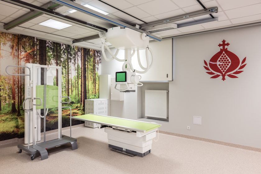 Zdjęcie przedstawia wnętrze nowoczesnej Pracowni Diagnostyki Obrazowej po zakończonej modernizacji. W centrum znajduje się cyfrowy aparat RTG z zawieszeniem sufitowym oraz stół diagnostyczny z jasnozielonym blatem. Po lewej stronie ustawiono pionowy statyw do wykonywania badań w pozycji stojącej, a ścianę zdobi fototapeta przedstawiająca słoneczny las. Po prawej stronie widoczne jest duże czerwone logo Zakonu Bonifratrów umieszczone na jasnoszarej ścianie. Jasne, przestronne wnętrze podkreśla nowoczesny charakter pracowni i wysoki standard wyposażenia.