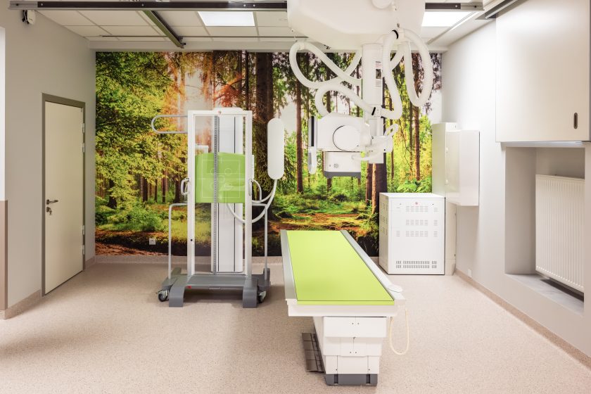 Zdjęcie przedstawia nowoczesną Pracownię Diagnostyki Obrazowej wyposażoną w cyfrowy aparat RTG z zawieszeniem sufitowym. Na pierwszym planie znajduje się stół diagnostyczny z charakterystycznym jasnozielonym blatem oraz pionowy statyw do wykonywania zdjęć rentgenowskich. W tle ścianę zdobi wielkoformatowa fototapeta przedstawiająca słoneczny las, nadająca wnętrzu przyjazny charakter. Pomieszczenie jest przestronne, jasne i utrzymane w nowoczesnej stylistyce, z jasną podłogą, zabudową techniczną oraz urządzeniami niezbędnymi do wykonywania badań radiologicznych.