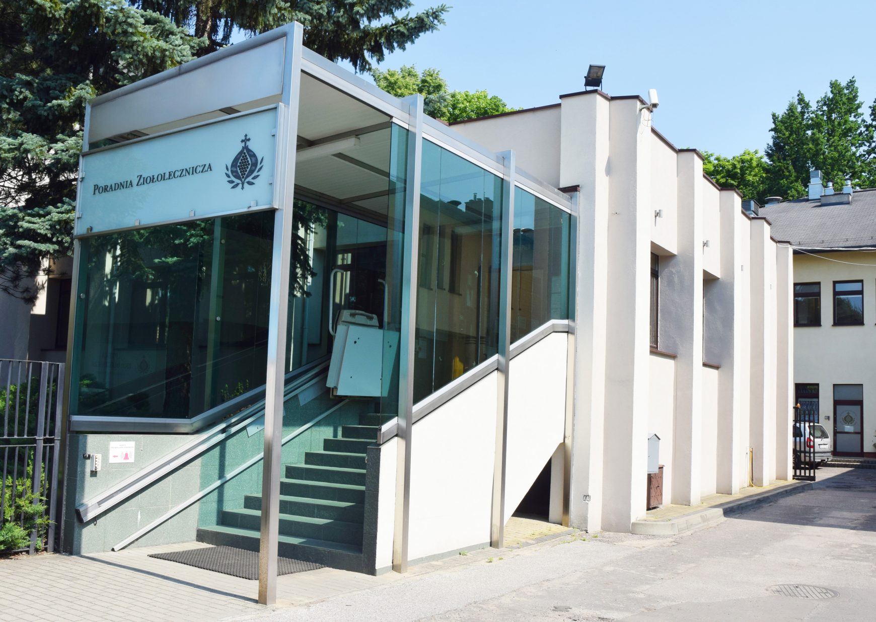 Zdjęcie obrazuje wejście do Poradni Ziołoleczniczej Bonifraterskiego Centrum Medycznego. Na pierwszym planie znajduje się nowoczesna, przeszklona zabudowa osłaniająca schody oraz platformę dla osób z niepełnosprawnościami, prowadzące do wejścia do budynku. Na górnym panelu widoczny jest napis „Poradnia Ziołolecznicza” oraz logo Bonifratrów. Po prawej stronie rozciąga się jasna elewacja budynku z charakterystycznymi pionowymi filarami i oknami. W tle widoczne są wysokie drzewa oraz bezchmurne, błękitne niebo. Zdjęcie wykonano w słoneczny dzień, a całość podkreśla zadbane i dostępne dla pacjentów wejście do placówki medycznej.