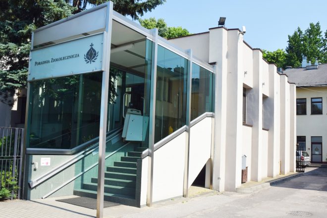 Zdjęcie obrazuje wejście do Poradni Ziołoleczniczej Bonifraterskiego Centrum Medycznego. Na pierwszym planie znajduje się nowoczesna, przeszklona zabudowa osłaniająca schody oraz platformę dla osób z niepełnosprawnościami, prowadzące do wejścia do budynku. Na górnym panelu widoczny jest napis „Poradnia Ziołolecznicza” oraz logo Bonifratrów. Po prawej stronie rozciąga się jasna elewacja budynku z charakterystycznymi pionowymi filarami i oknami. W tle widoczne są wysokie drzewa oraz bezchmurne, błękitne niebo. Zdjęcie wykonano w słoneczny dzień, a całość podkreśla zadbane i dostępne dla pacjentów wejście do placówki medycznej.