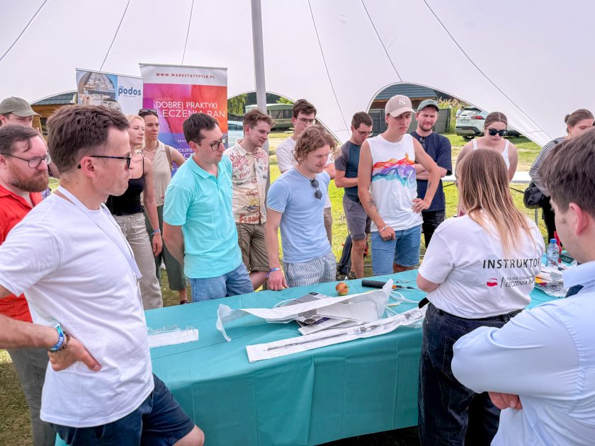 Zdjęcie obrazuje grupę uczestników zgromadzonych wokół stanowiska warsztatowego podczas praktycznych zajęć z leczenia ran. Instruktorka, stojąca tyłem do obiektywu, prezentuje sprzęt medyczny zgromadzony na stole. Wokół niej stoi kilkanaście osób uważnie obserwujących pokaz. W tle widoczne są roll-upy Polskiego Towarzystwa Leczenia Ran oraz Warsztatów Dobrej Praktyki Leczenia Ran.