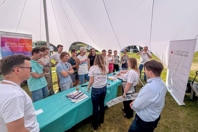 Zdjęcie obrazuje szeroki plan warsztatów praktycznych prowadzonych pod dużym białym namiotem podczas konferencji ChiCamp 2026. Dwie instruktorki Polskiego Towarzystwa Leczenia Ran prowadzą pokaz przy stole wypełnionym narzędziami i materiałami medycznymi. Wokół stanowiska stoi liczna grupa uczestników, a po prawej stronie widoczny jest prelegent obserwujący przebieg zajęć. Po bokach ustawiono roll-upy Polskiego Towarzystwa Leczenia Ran oraz Warsztatów Dobrej Praktyki Leczenia Ran, podkreślające charakter edukacyjny wydarzenia.