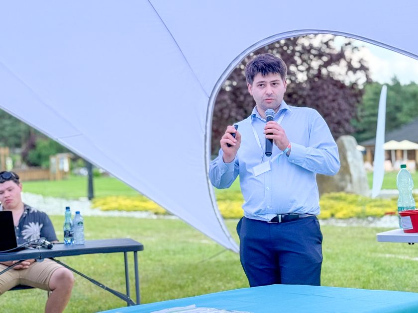 Zdjęcie obrazuje wykład prowadzony podczas konferencji ChiCamp 2026. Mężczyzna ubrany w jasnoniebieską koszulę stoi pod dużym namiotem i przemawia do uczestników, trzymając w jednej dłoni mikrofon, a w drugiej pilot do prezentacji. W tle widoczny jest zielony teren z drzewami oraz stolik z komputerem obsługującym prezentację multimedialną.