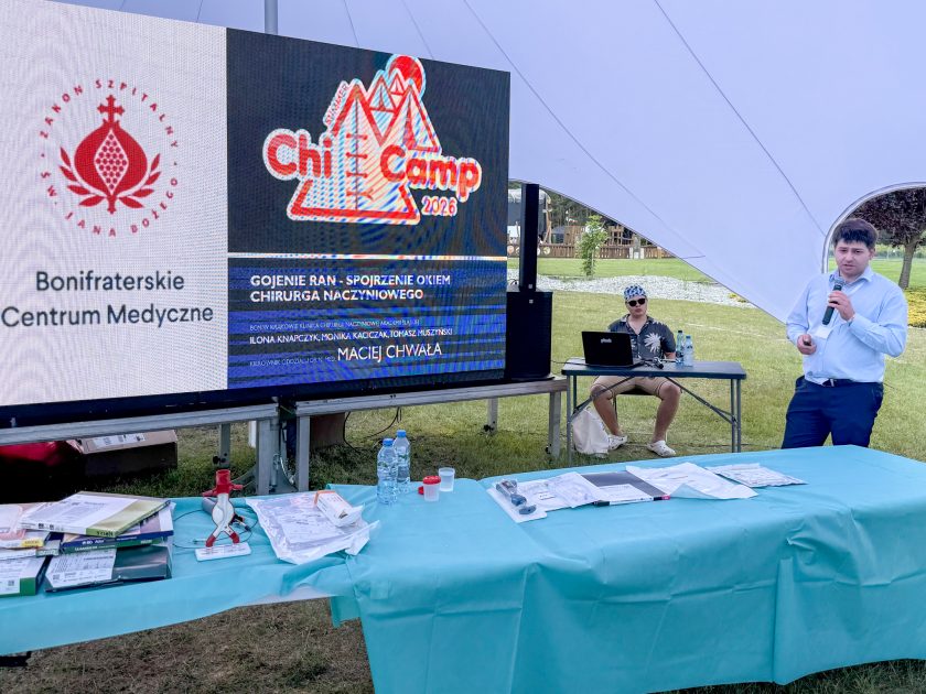 Zdjęcie obrazuje prezentację pt. „Gojenie ran – spojrzenie okiem chirurga naczyniowego” prowadzoną podczas konferencji ChiCamp 2026. Po lewej stronie znajduje się duży ekran LED z logotypami Bonifraterskiego Centrum Medycznego, Zakonu Szpitalnego św. Jana Bożego oraz wydarzenia ChiCamp 2026, a także tytułem wykładu i nazwiskami prowadzących. Po prawej stronie stoi prelegent z mikrofonem. Na pierwszym planie widoczny jest stół przygotowany do warsztatów z narzędziami i materiałami medycznymi.