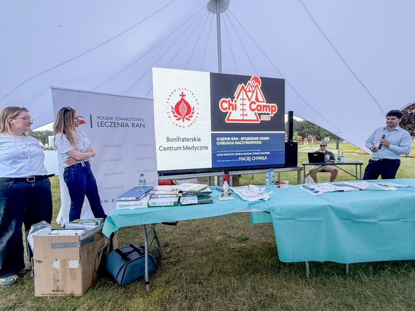Zdjęcie obrazuje początek warsztatów praktycznych prowadzonych podczas ChiCamp 2026. Po lewej stronie stoją dwie instruktorki w koszulkach Polskiego Towarzystwa Leczenia Ran, a po prawej prelegent z mikrofonem. Centralną część zdjęcia zajmuje duży ekran LED z prezentacją dotyczącą leczenia ran z perspektywy chirurga naczyniowego oraz logotypami Bonifraterskiego Centrum Medycznego i Zakonu Szpitalnego św. Jana Bożego. Na stołach rozmieszczono zestawy sprzętu medycznego wykorzystywanego podczas ćwiczeń.