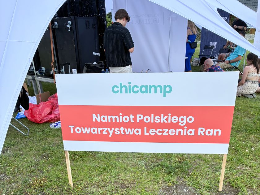 Zdjęcie obrazuje tablicę informacyjną ustawioną przed namiotem warsztatowym. Na białym tle znajduje się zielony napis „chicamp”, a poniżej czerwony pas z dużym białym napisem „Namiot Polskiego Towarzystwa Leczenia Ran”. W tle widoczne są elementy zaplecza technicznego wydarzenia oraz uczestnicy przebywający pod namiotem.