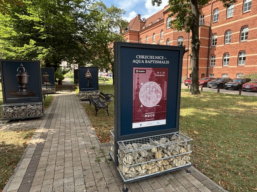 Plenerowa wystawa „Chrzcielnice – Aqua Baptismalis” ustawiona w ogrodzie przy zabytkowym budynku z czerwonej cegły. Na pierwszym planie znajduje się tablica z tytułem wystawy oraz plakatem krakowskiego wydarzenia „Noc Cracovia Sacra”, a wzdłuż alejki stoją kolejne plansze prezentujące fotografie zabytkowych chrzcielnic. Tablice umieszczono na metalowych konstrukcjach wypełnionych kamieniami.
