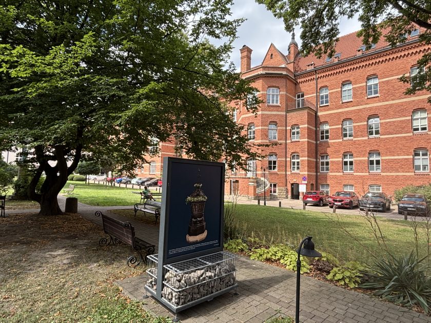 Jedna z plansz plenerowej wystawy poświęconej zabytkowym chrzcielnicom, ustawiona przy alejce w zielonym ogrodzie. Na planszy widoczna jest fotografia bogato zdobionej chrzcielnicy. W tle znajduje się rozłożyste drzewo, ławki oraz wielokondygnacyjny, zabytkowy budynek z czerwonej cegły.