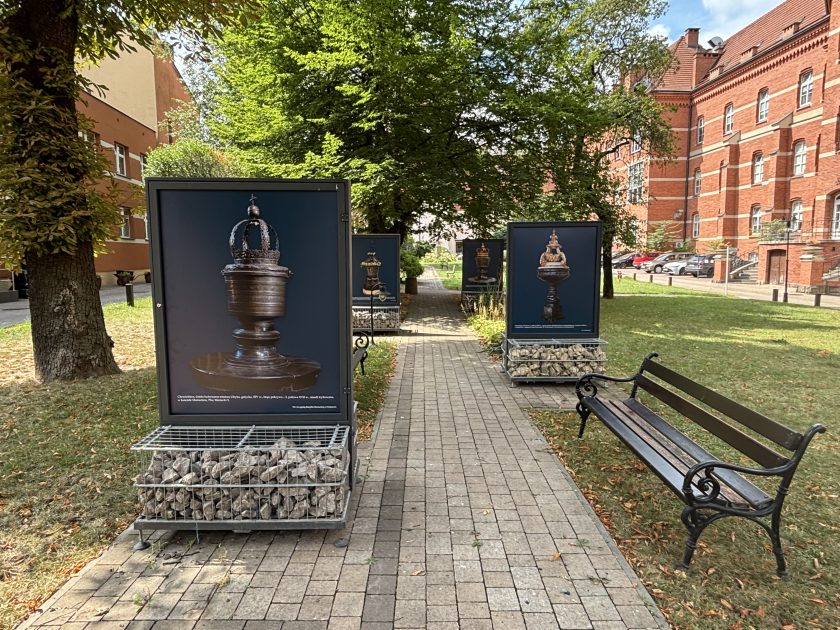 Widok wzdłuż alejki z plenerową wystawą prezentującą fotografie zabytkowych chrzcielnic. Po obu stronach chodnika ustawiono duże plansze na metalowych podstawach wypełnionych kamieniami. Na pierwszym planie widoczna jest fotografia okazałej, metalowej chrzcielnicy zwieńczonej koroną. Po prawej stronie stoi drewniana ławka, a w tle widać drzewa i zabytkowy budynek z czerwonej cegły.