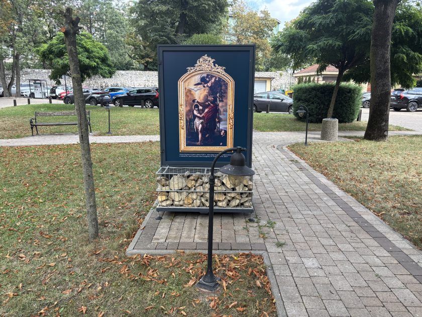 Plansza plenerowej wystawy ustawiona na ogrodowej alejce, przedstawiająca obraz sceny chrztu Jezusa w Jordanie, oprawiony w ozdobną złotą ramę. Plansza stoi na metalowej podstawie wypełnionej kamieniami. Wokół znajdują się drzewa, trawnik i ławki, a w tle widoczny jest parking oraz kamienny mur.