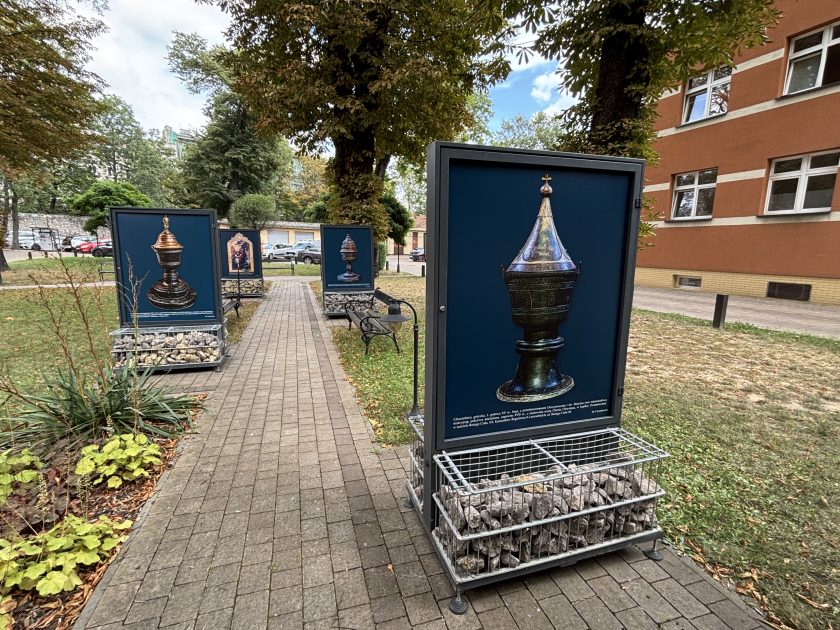 Plenerowa wystawa fotografii zabytkowych chrzcielnic rozmieszczona wzdłuż alejki w ogrodzie. Na pierwszym planie znajduje się plansza ze zdjęciem wysokiej, metalowej chrzcielnicy ze stożkowatą, bogato zdobioną pokrywą zwieńczoną krzyżem. W głębi widoczne są kolejne plansze przedstawiające chrzcielnice oraz obraz o tematyce chrztu Jezusa.