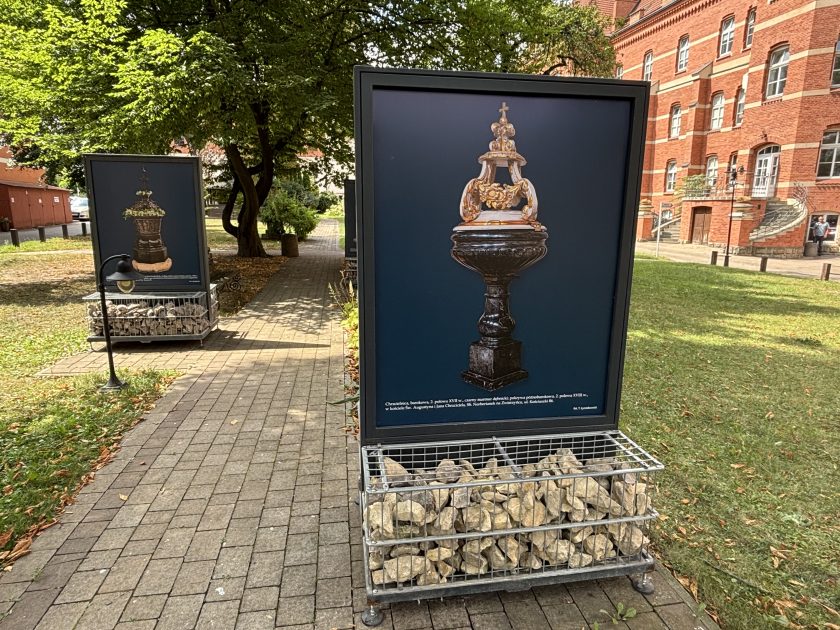 Plansza plenerowej wystawy prezentująca fotografię zabytkowej chrzcielnicy z ciemną kamienną czaszą i bogato zdobioną, jasną pokrywą zwieńczoną krzyżem. Plansza ustawiona jest na metalowej podstawie wypełnionej kamieniami przy ogrodowej alejce. W tle widoczna jest kolejna plansza wystawowa, drzewa oraz zabytkowy budynek z czerwonej cegły.