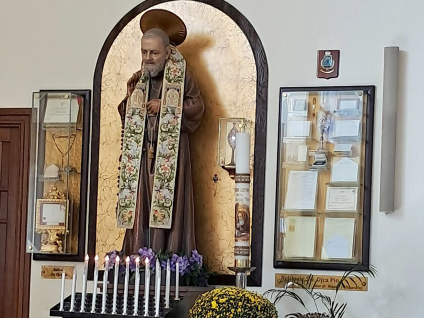 Figura św. Ojca Pio we wnętrzu sanktuarium, otoczona świecami, kwiatami oraz gablotami z pamiątkami.