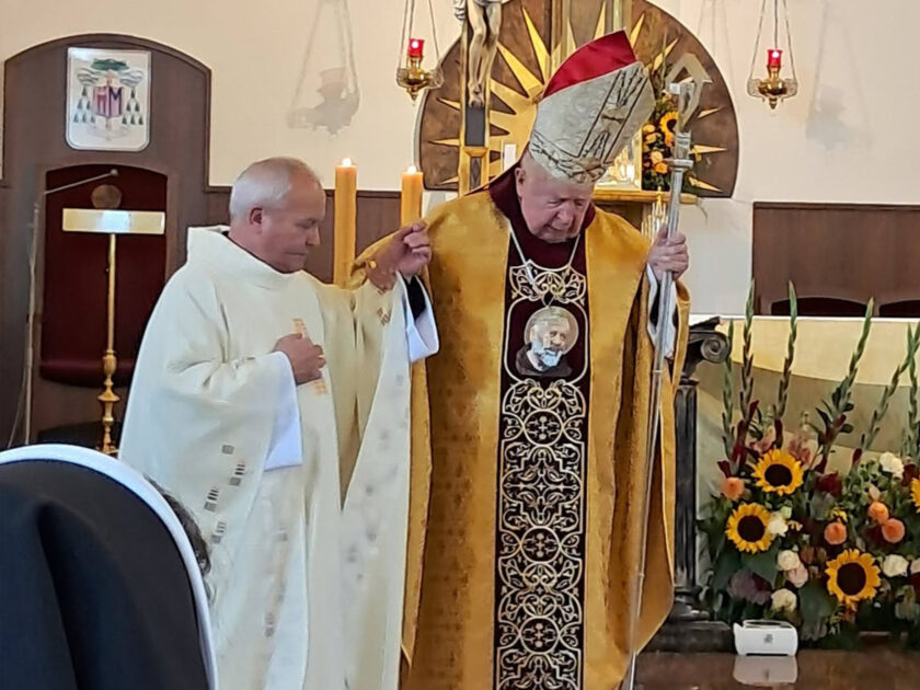 Biskup w szatach liturgicznych wraz z duchownym podczas uroczystości przy ołtarzu. W tle widoczny krucyfiks oraz religijny wizerunek Chrystusa.