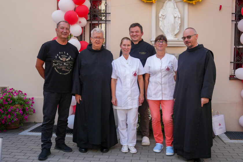 Pamiątkowe zdjęcie sześciu osób podczas jubileuszu warszawskich dzieł bonifraterskich. W grupie znajdują się dwaj bracia zakonni w czarnych habitach, dwie kobiety w białych strojach medycznych oraz dwóch mężczyzn. W tle widoczna jest figura Matki Bożej oraz biało-czerwone balony zdobiące budynek.