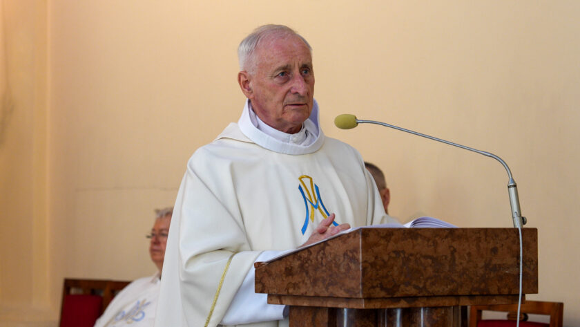 Starszy kapłan w białych szatach liturgicznych stoi przy ambonie i przemawia podczas Mszy Świętej odprawianej w ramach jubileuszu warszawskich dzieł bonifraterskich. Przed nim znajduje się mikrofon i otwarta księga liturgiczna.