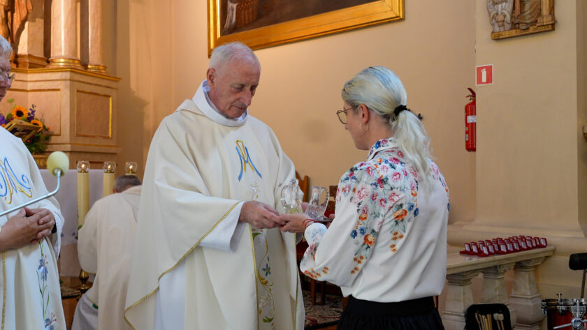 Kapłan w białych szatach liturgicznych przekazuje kobiecie niewielkie szklane upominki podczas uroczystości jubileuszowej w kościele. W tle widoczny jest ołtarz, świece oraz drugi duchowny.