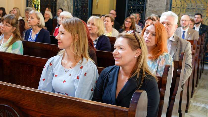 Uczestnicy jubileuszowej Mszy Świętej siedzą w drewnianych ławkach kościoła i słuchają trwającej uroczystości. Na pierwszym planie widoczne są trzy kobiety, a za nimi kolejni uczestnicy spotkania.