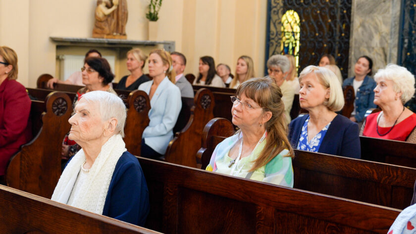 Uczestnicy jubileuszowej Mszy Świętej zgromadzeni w ławkach kościoła. Na pierwszym planie siedzi starsza kobieta w granatowym stroju i białym szalu, a za nią kolejne osoby skupione na przebiegu nabożeństwa.