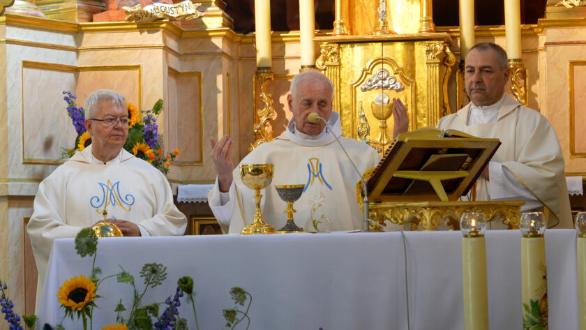 Trzech duchownych w białych szatach liturgicznych przy ołtarzu podczas sprawowania jubileuszowej Mszy Świętej. Kapłan stojący pośrodku unosi dłonie w geście modlitwy, a przed nim na ołtarzu znajdują się kielichy. W tle widoczny jest bogato złocony ołtarz.