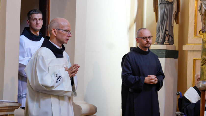 Bracia zakonni uczestniczący w jubileuszowej Mszy Świętej. Na pierwszym planie zakonnik w białej szacie liturgicznej z czarnym kapturem stoi ze złożonymi do modlitwy dłońmi, obok niego brat w czarnym habicie. W tle widoczny jest kolejny uczestnik liturgii oraz fragment wnętrza kościoła.
