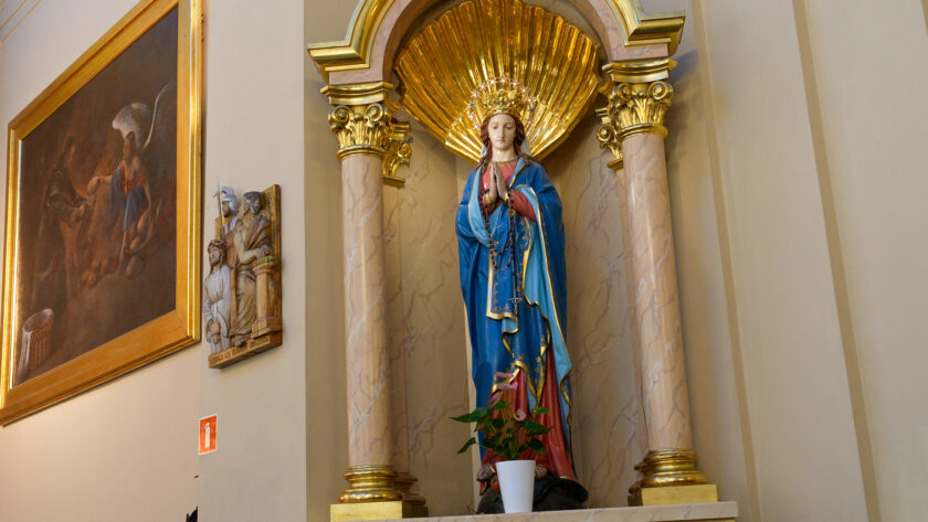 Figura Matki Bożej w niebiesko-białych szatach, umieszczona w bogato zdobionej, złoconej niszy we wnętrzu kościoła. Po lewej stronie widoczny jest duży obraz religijny oraz niewielka płaskorzeźba przedstawiająca scenę biblijną.