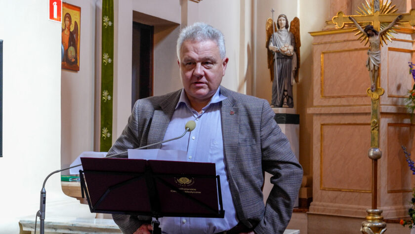 Mężczyzna w jasnoniebieskiej koszuli i szarej marynarce w kratę przemawia do mikrofonu podczas jubileuszowego spotkania w kościele. Przed nim znajduje się pulpit z materiałami Bonifraterskiego Centrum Medycznego. W tle widoczne są elementy wyposażenia świątyni, figura anioła oraz krucyfiks.