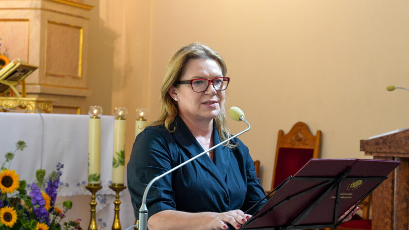 Kobieta w ciemnej sukience i czerwonych okularach przemawia przy mikrofonie podczas uroczystości jubileuszowej w kościele. Stoi przy pulpicie z oznaczeniem Bonifraterskiego Centrum Medycznego. W tle widoczny jest ołtarz ozdobiony świecami oraz kompozycją kwiatową ze słonecznikami i fioletowymi kwiatami.