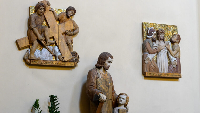 Fragment wystroju kościoła z drewnianymi płaskorzeźbami religijnymi. Po lewej znajduje się stacja Drogi Krzyżowej z napisem „Trzeci raz Jezus na drodze upada”, po prawej scena „Jezus z szat swoich obnażony”. Pomiędzy nimi stoi drewniana figura świętego z dzieckiem.