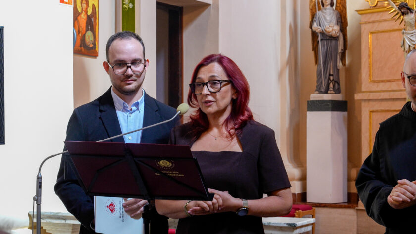 Kobieta o rudych włosach i w okularach przemawia do mikrofonu podczas jubileuszowej uroczystości w kościele. Obok niej stoi mężczyzna w ciemnej marynarce, trzymający teczkę z logo Bonifraterskiego Centrum Medycznego. Z prawej strony kadru widoczny jest fragment brata zakonnego w czarnym habicie.