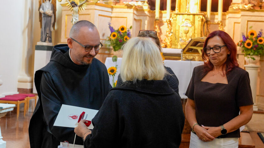 Moment wręczania podziękowania podczas jubileuszu warszawskich dzieł bonifraterskich. Brat zakonny w czarnym habicie z uśmiechem przyjmuje od stojącej tyłem kobiety pamiątkowy upominek. Obok stoi rudowłosa kobieta w ciemnym stroju. W tle widoczny jest bogato zdobiony ołtarz oraz kompozycje ze słoneczników i innych kwiatów.